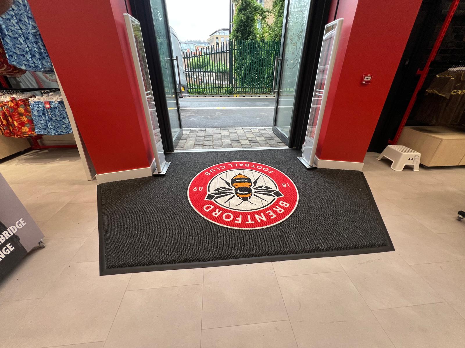 Entrance to Brentford FC Fanstore showing a grey precision nib logo mat with the Brentford FC Emblem designed and made by COBA Flooring.
