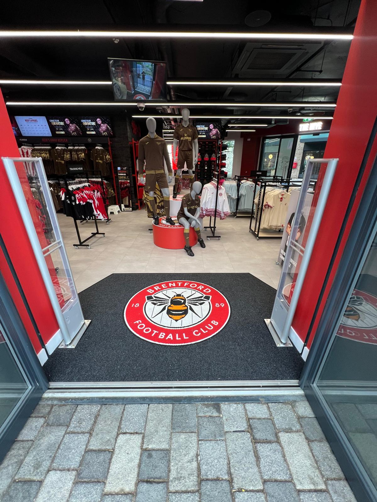 Entrance to Brentford FC Fanstore showing a grey precision nib logo mat with the Brentford FC Emblem designed and made by COBA Flooring.