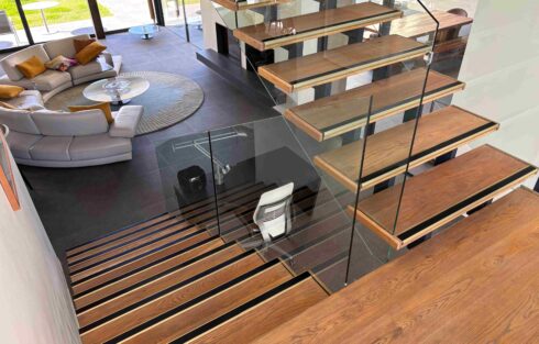 Modern floating wooden staircase with glass balustrades and black anti slip stair inserts overlooking a contemporary living area.